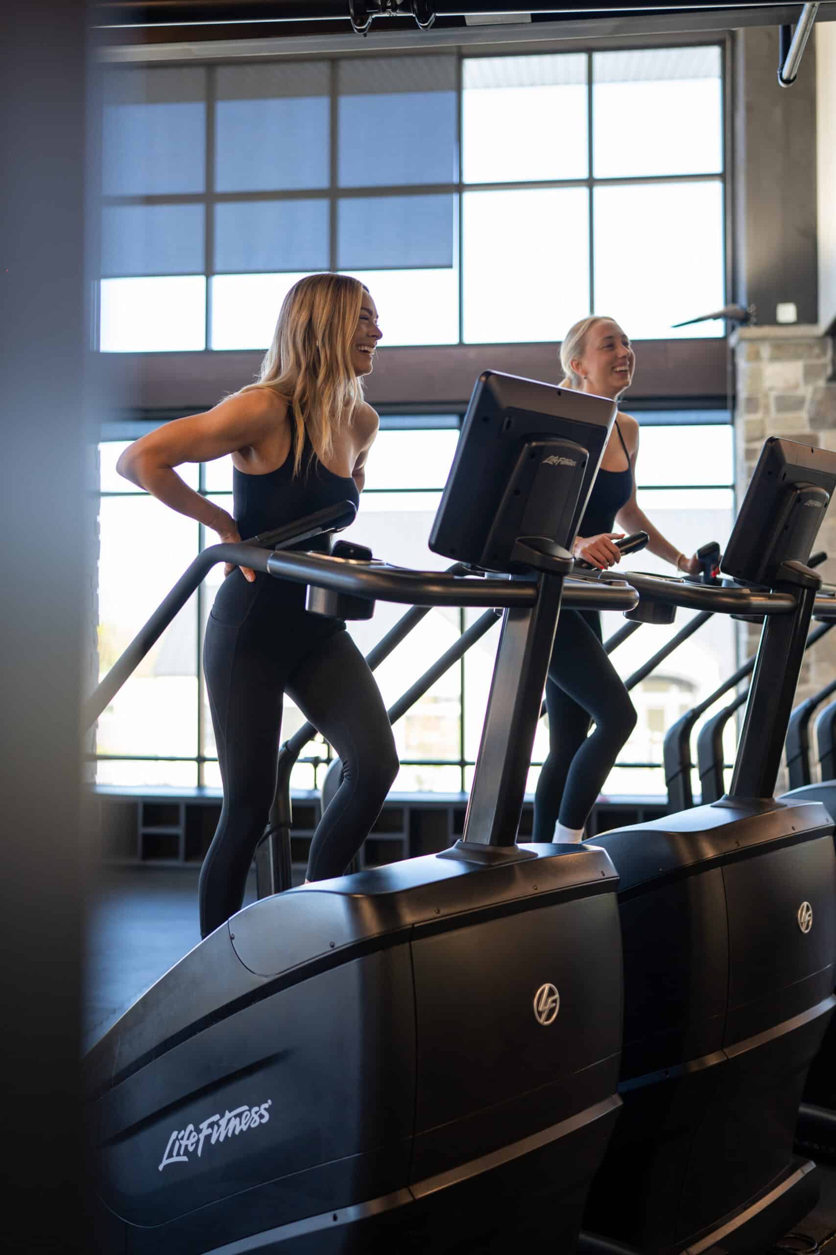 Two smiling women working out on Stairmasters at Alpine Fitness, a gym in American Fork, UT offering personal training, nutrition consulting, cardio and strength training, cold plunge and sauna therapy, massage therapy, and muscle recovery programs. Reach out to us now for help with gym in American Fork UT, gym near American Fork UT, fitness center in American Fork UT, local gym in American Fork Utah, best gym in American Fork UT, gym near me in American Fork, personal training in American Fork UT, personal trainer near me, certified personal trainer American Fork UT, nutrition consulting American Fork UT, nutrition coach near me, performance support programs American Fork UT, strength and conditioning near me, weight loss programs in American Fork UT, cardio training gym American Fork UT, functional fitness programs near me, athletic performance training American Fork UT, post-rehabilitation programs near me, recovery gym American Fork UT, muscle recovery therapy near me, massage therapy American Fork UT, massage near me, cold plunge therapy American Fork UT, sauna therapy near me, gym with recovery services in American Fork UT, small group training programs near me, one-on-one personal training American Fork UT, personalized fitness programs near me, clean gym in American Fork UT, modern gym equipment near me, injury rehab programs American Fork UT, strength, cardio & weight loss training near me.